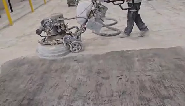 A worker pushing a walk-behind grinder across a large warehouse-style concrete floor, with a vacuum hose looping from the machine.