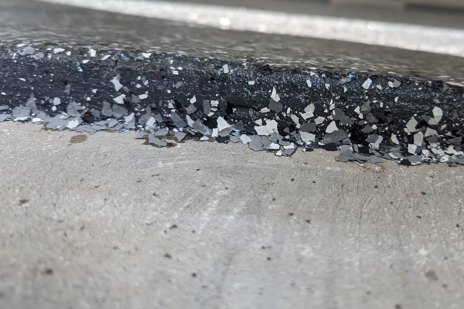 Close view of the cut edge of a finished flake floor beside bare concrete, showing vinyl flakes suspended in dark resin under a glossy clear surface.