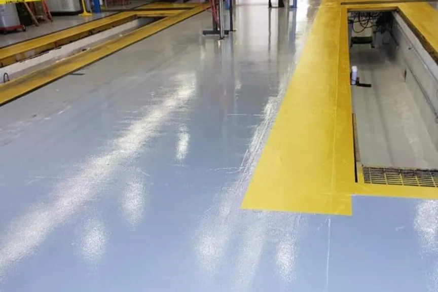 A coated floor in a vehicle service facility, light blue-gray with safety-yellow markings around the inspection pits.