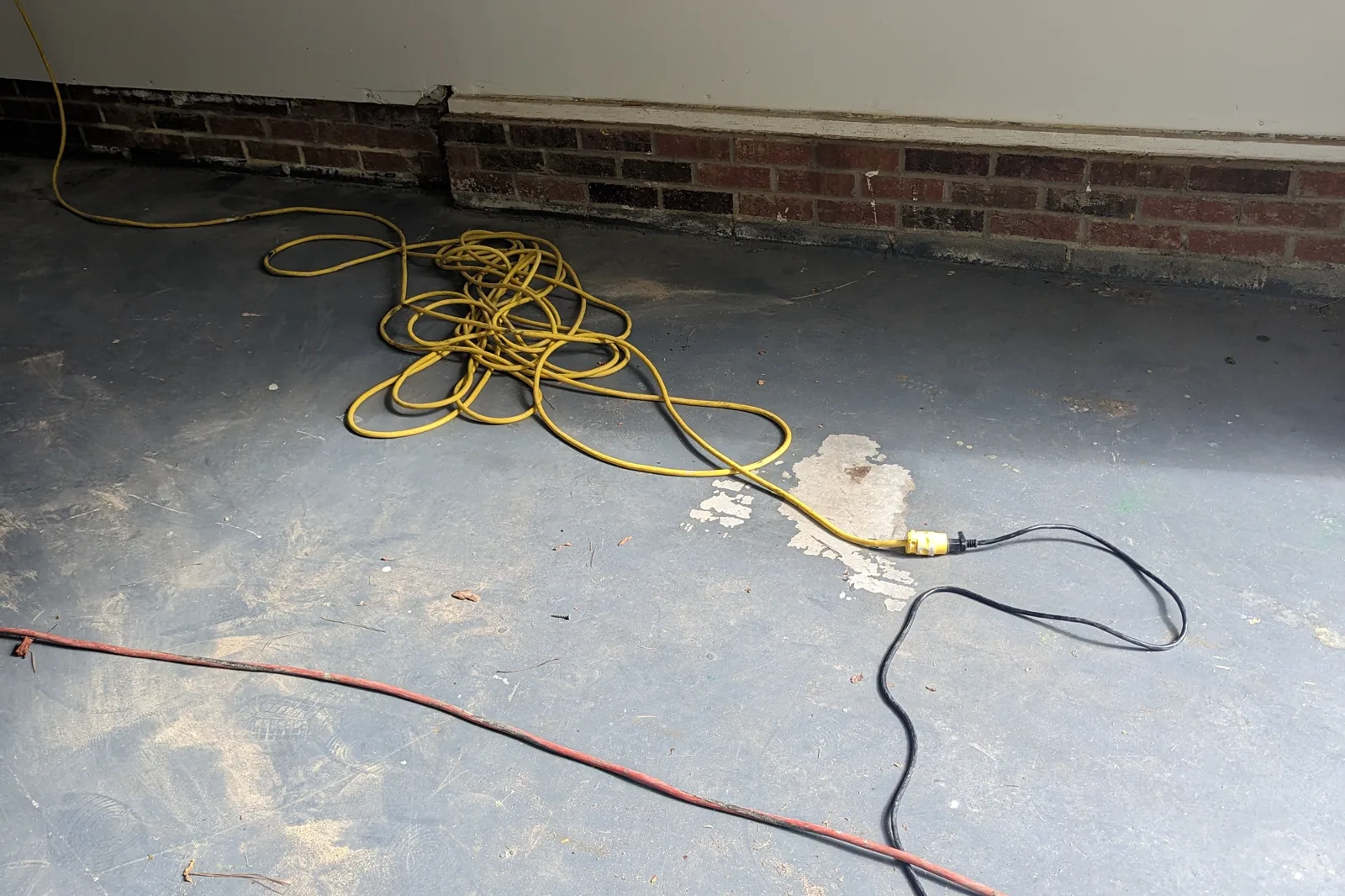 A garage floor with an existing gray coating in poor condition, worn through to bare concrete in patches, with extension cords across it and a brick knee wall along the side.