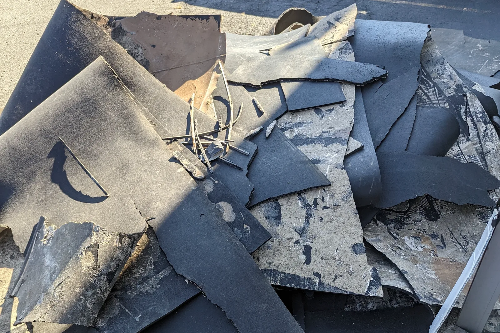 A pile of black sheet flooring torn out of a building, with adhesive and backing residue still attached to the pieces, stacked outside for disposal.