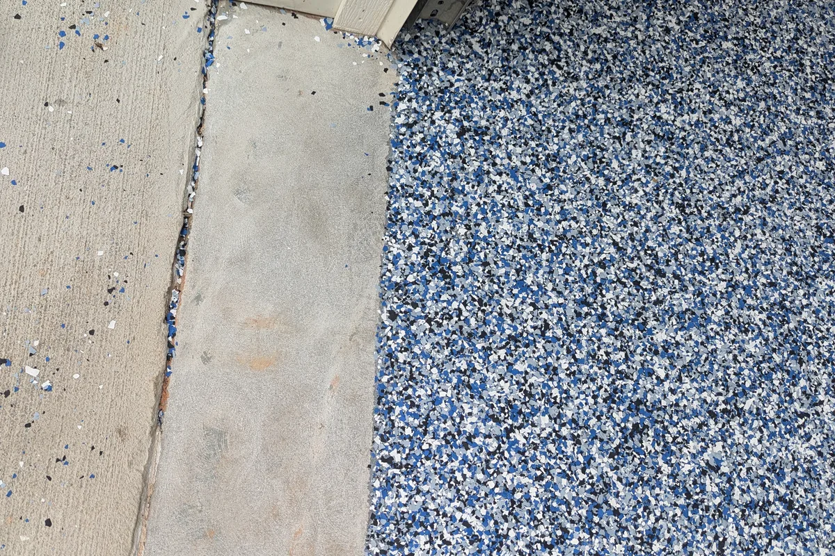 The edge of a finished flake floor at a garage threshold, with dense flake coverage on one side and loose stray flakes scattered across the bare concrete apron.