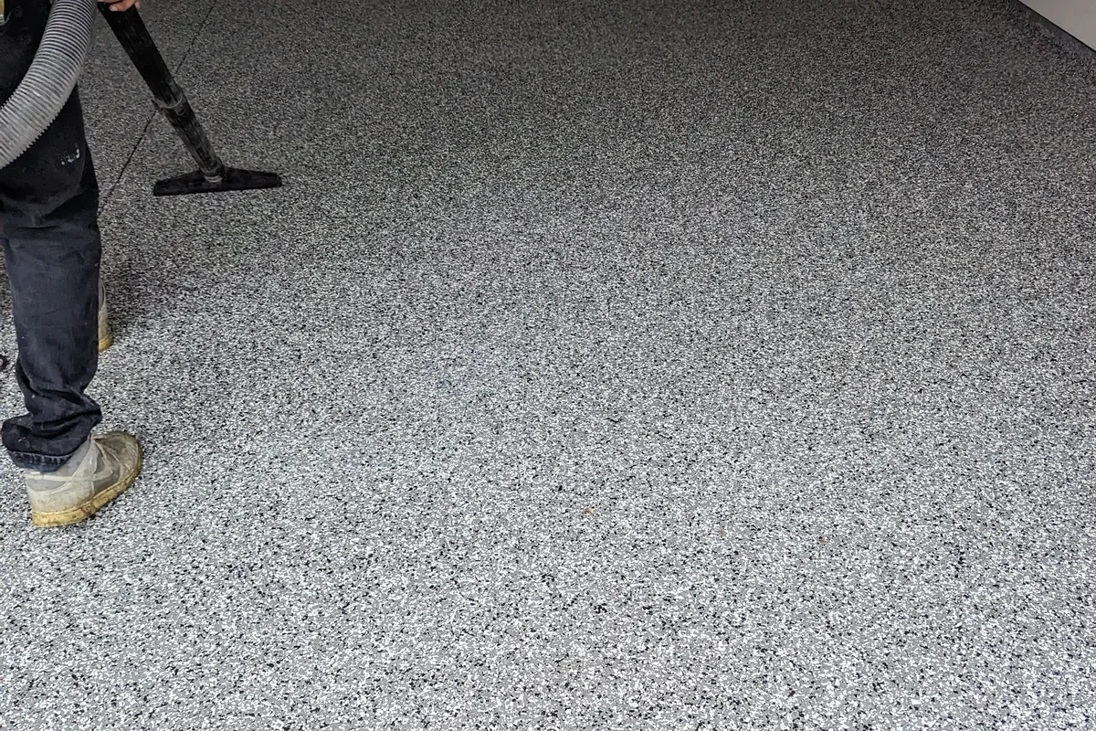 An installer using a vacuum to lift loose flake from a finished floor, standing on the coated surface in protective boot covers.