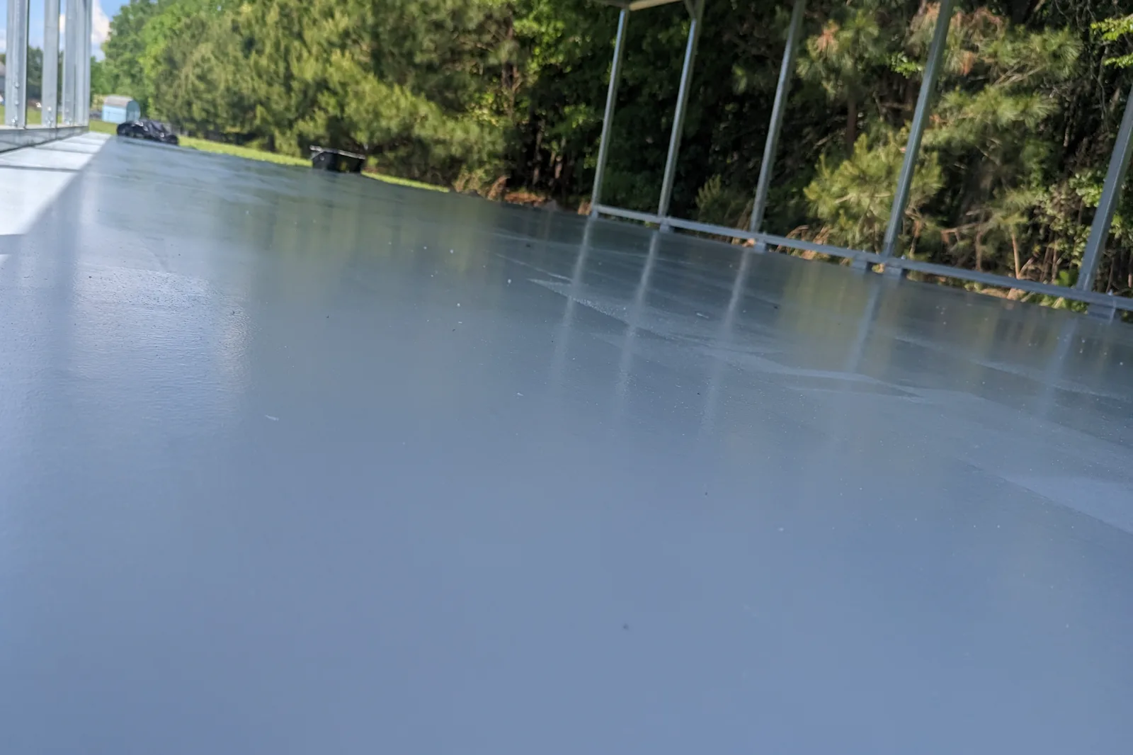 A solid gray coated slab under an open-sided steel canopy, sunlight falling across the surface with trees beyond.