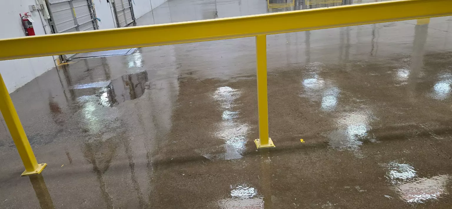 A commercial floor with a yellow safety railing, a fire extinguisher and loading doors in the background, wet and reflective during the sanding phase.