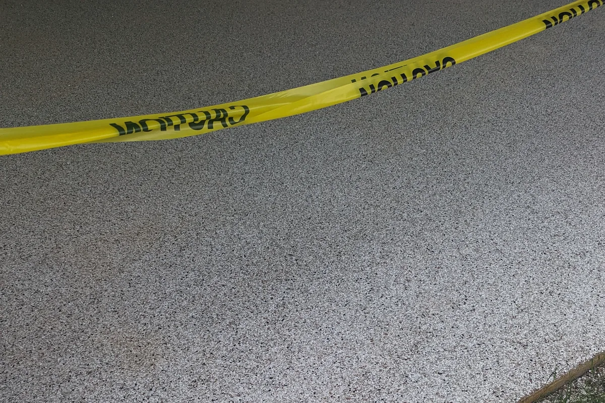 A newly finished exterior flake floor with caution tape across it, the coating running to a clean edge where it meets grass.
