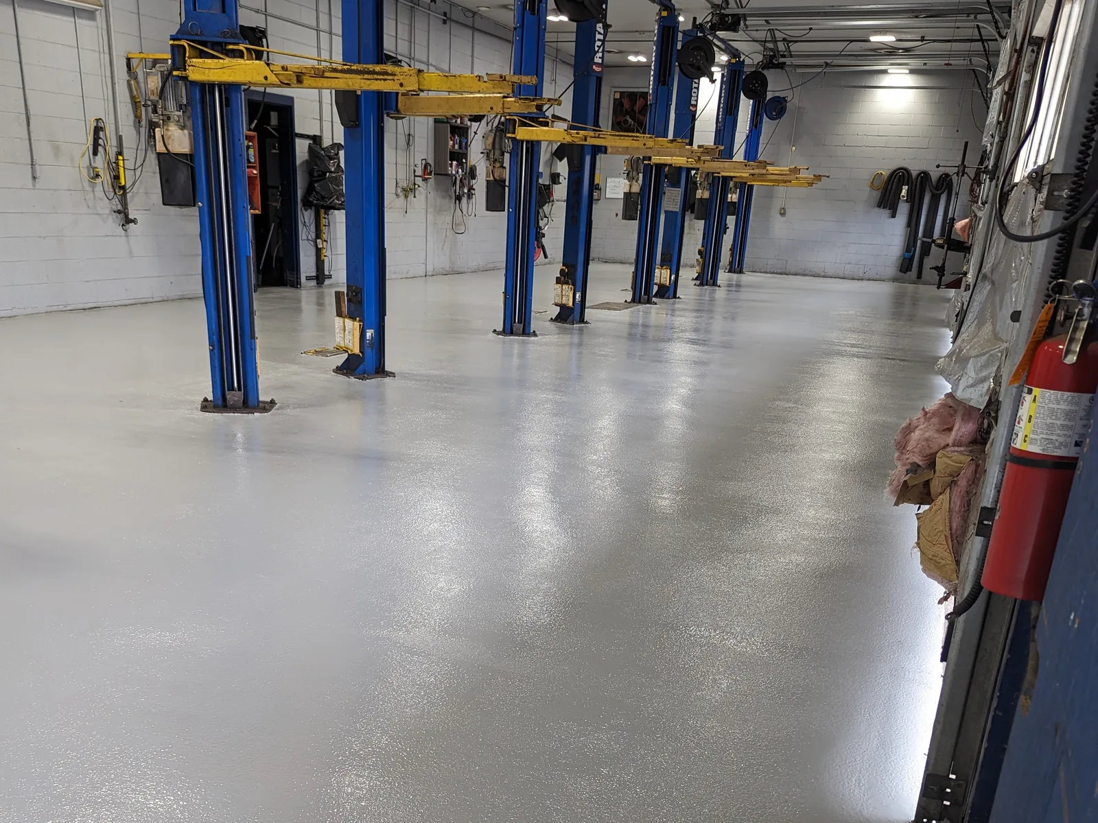 A long service bay with a smooth light grey coated floor, blue vehicle lifts standing along one side and a red fire extinguisher mounted on a post at the right.