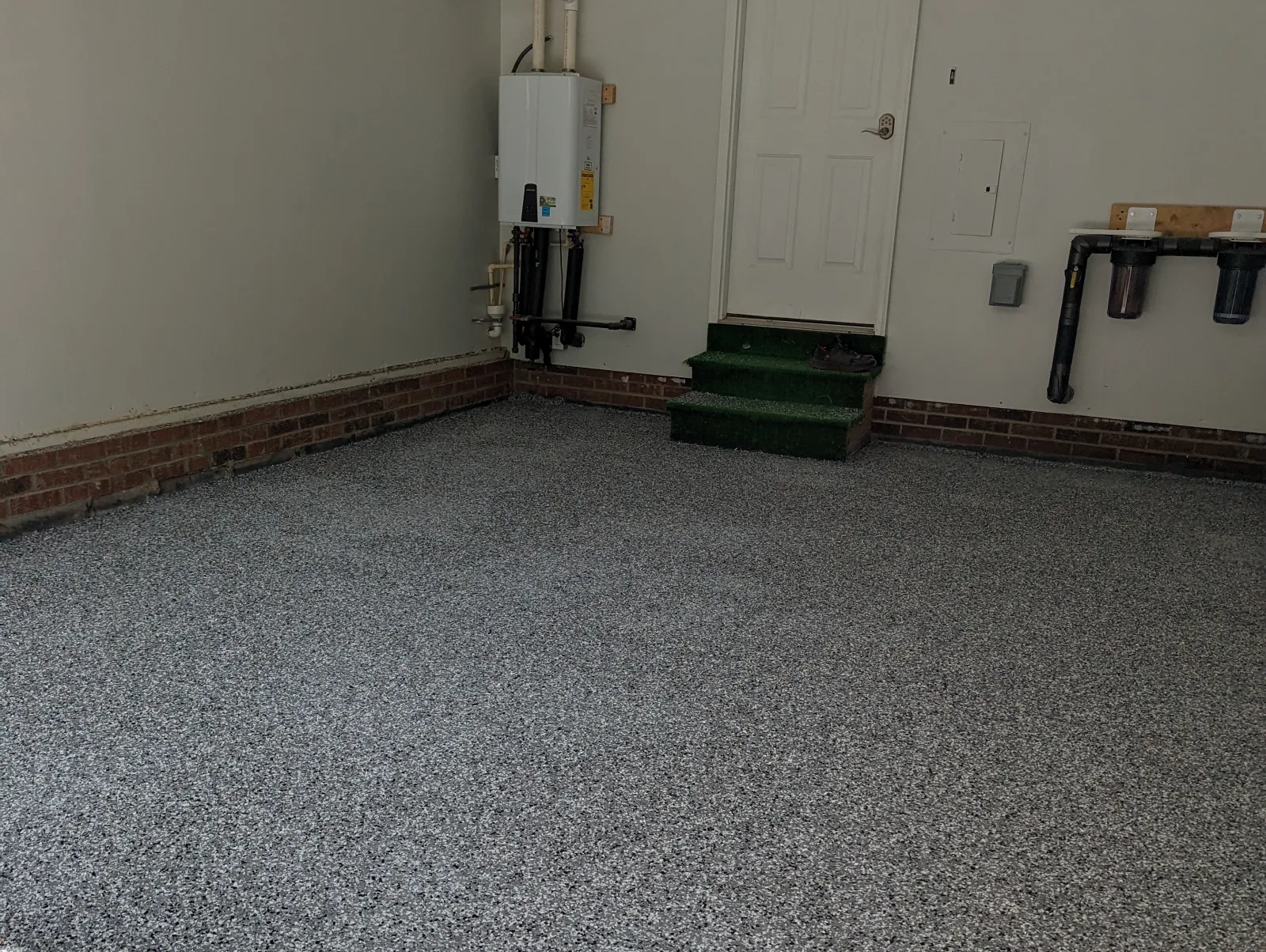 A garage floor finished in a gray, black and white flake blend, running to a white door with two green steps, a wall-mounted water heater and a brick base along the wall.