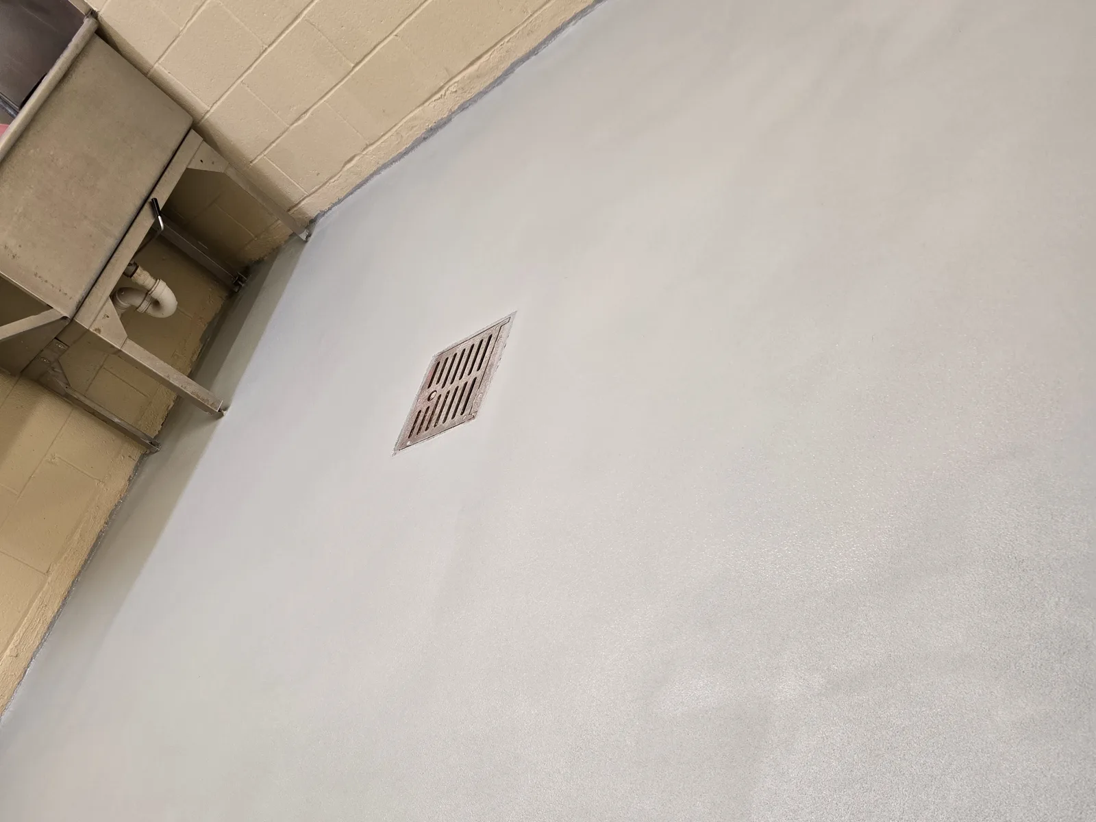 A floor drain grate set flush into a pale grey coated floor, with the coating turning up the wall as a coved base and stainless equipment legs standing at the left.