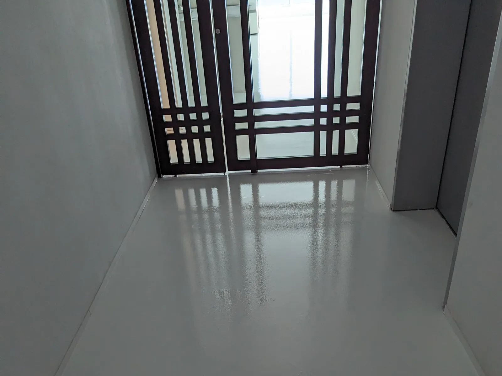 A commercial entry area with a smooth pale grey floor reflecting a set of dark-framed glass doors, with a second finished space visible beyond them.