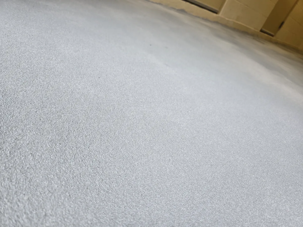 Close view of a pale grey floor surface with a fine granular texture, meeting a painted block wall where the coating turns up as a coved base.