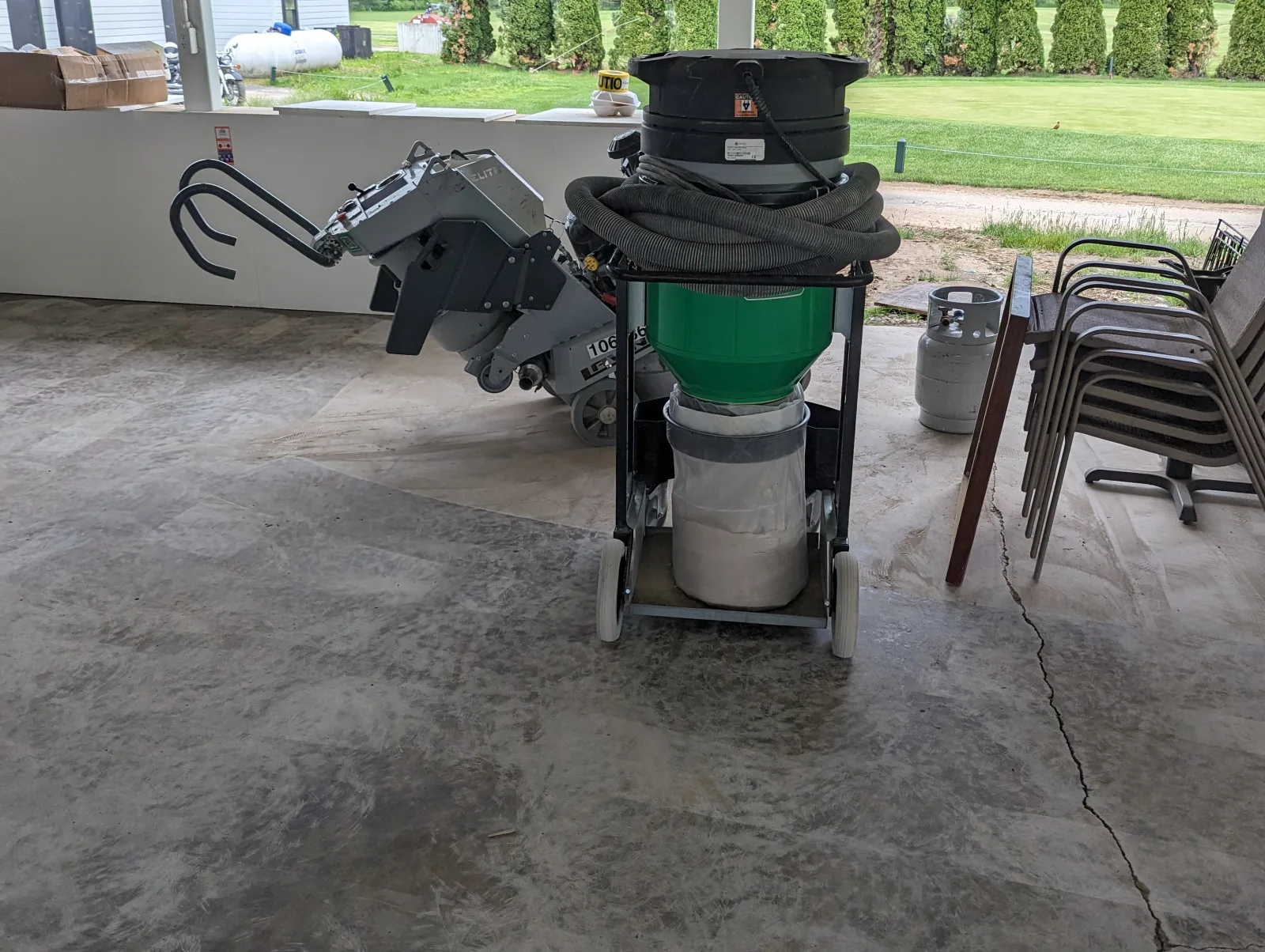 A floor grinder with its handles raised standing beside a wheeled dust extractor with a coiled hose, on a concrete slab under a roofed open-sided structure, with a crack running across the floor on the right.