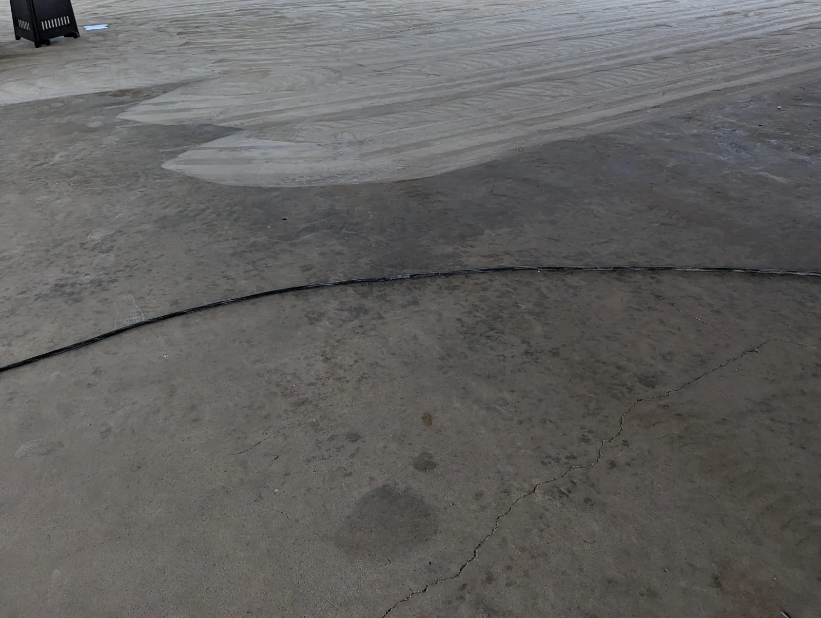A large concrete slab under a roof, with a pale rippled area of ground surface at the top, a dark line curving across the floor and a fine crack running through the darker concrete in the foreground.