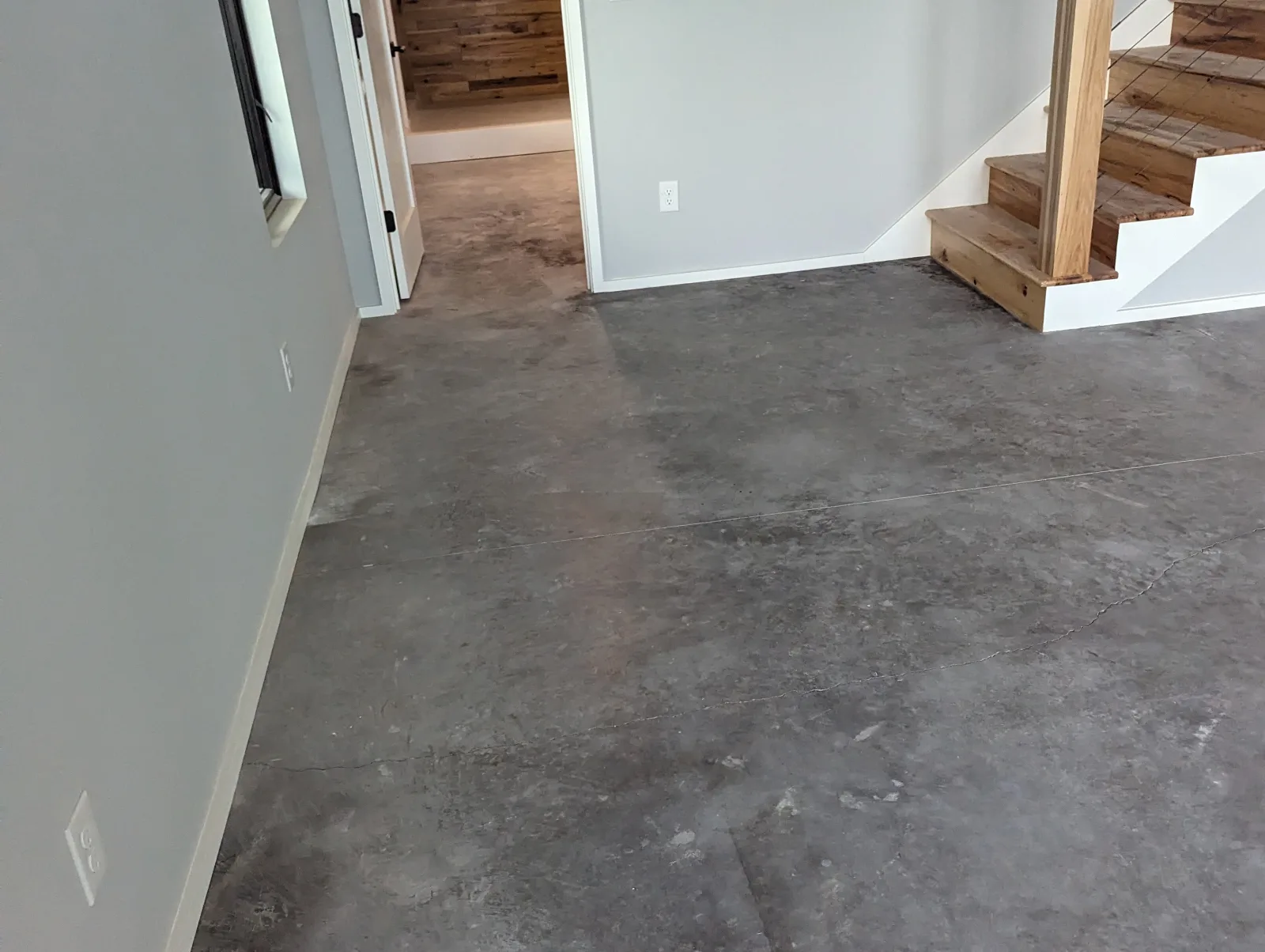A bare grey concrete floor with a mottled, uneven surface in an interior room, with a doorway on the left, a wooden staircase on the right and fine lines running across the slab.