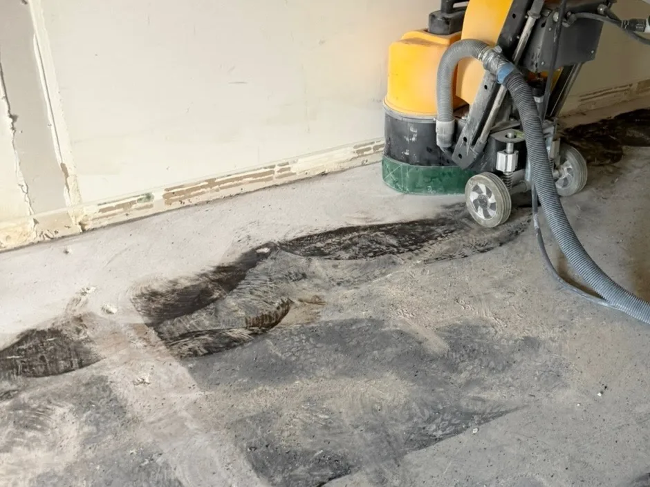 A walk-behind floor grinder with a dust extraction hose attached, working across a slab where dark existing floor material is partly removed and paler concrete is exposed around it.