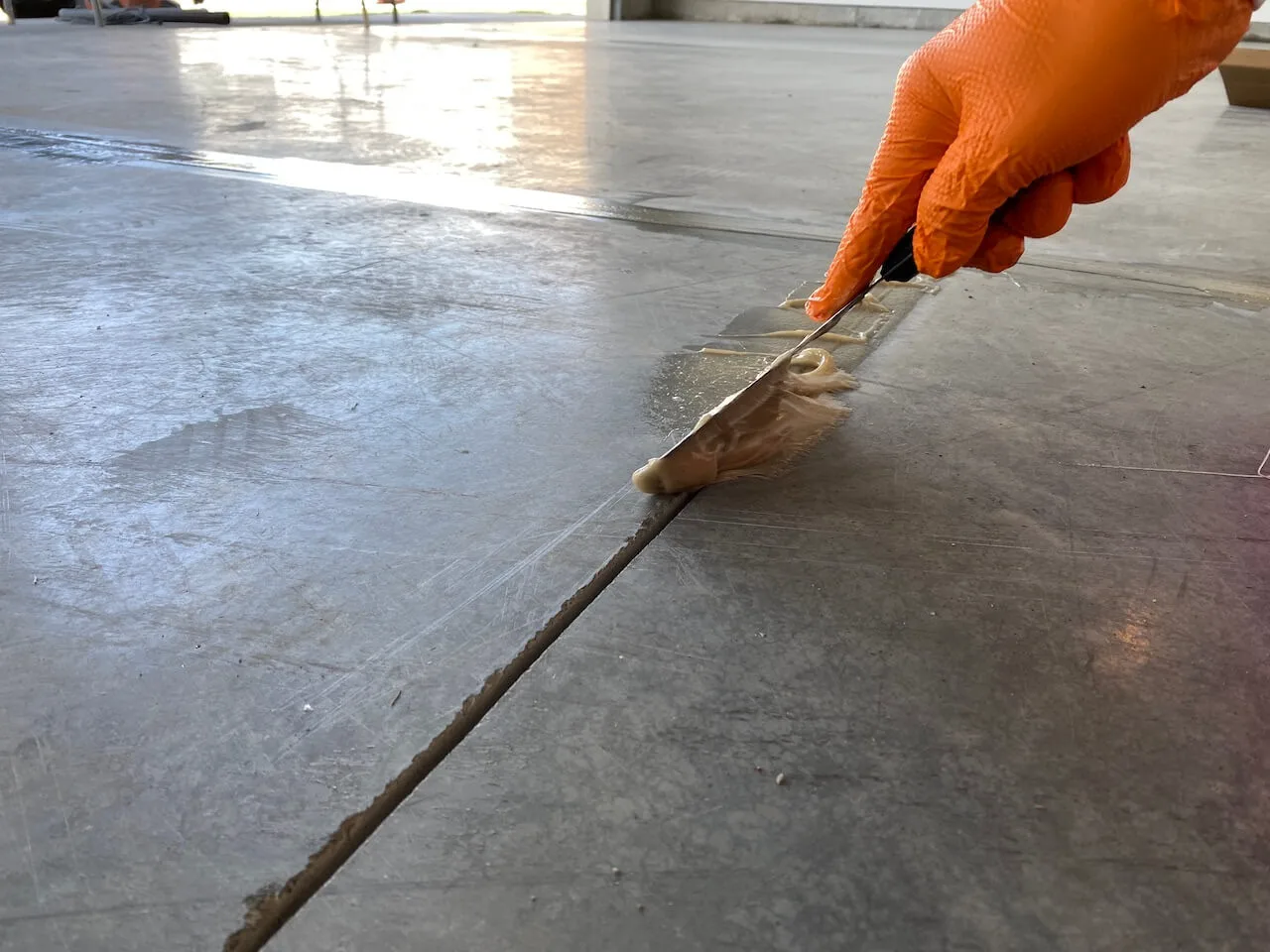 A gloved hand drawing a putty knife along a joint in a concrete slab, working a pale filler material into it, with grinder marks visible in the surrounding concrete.