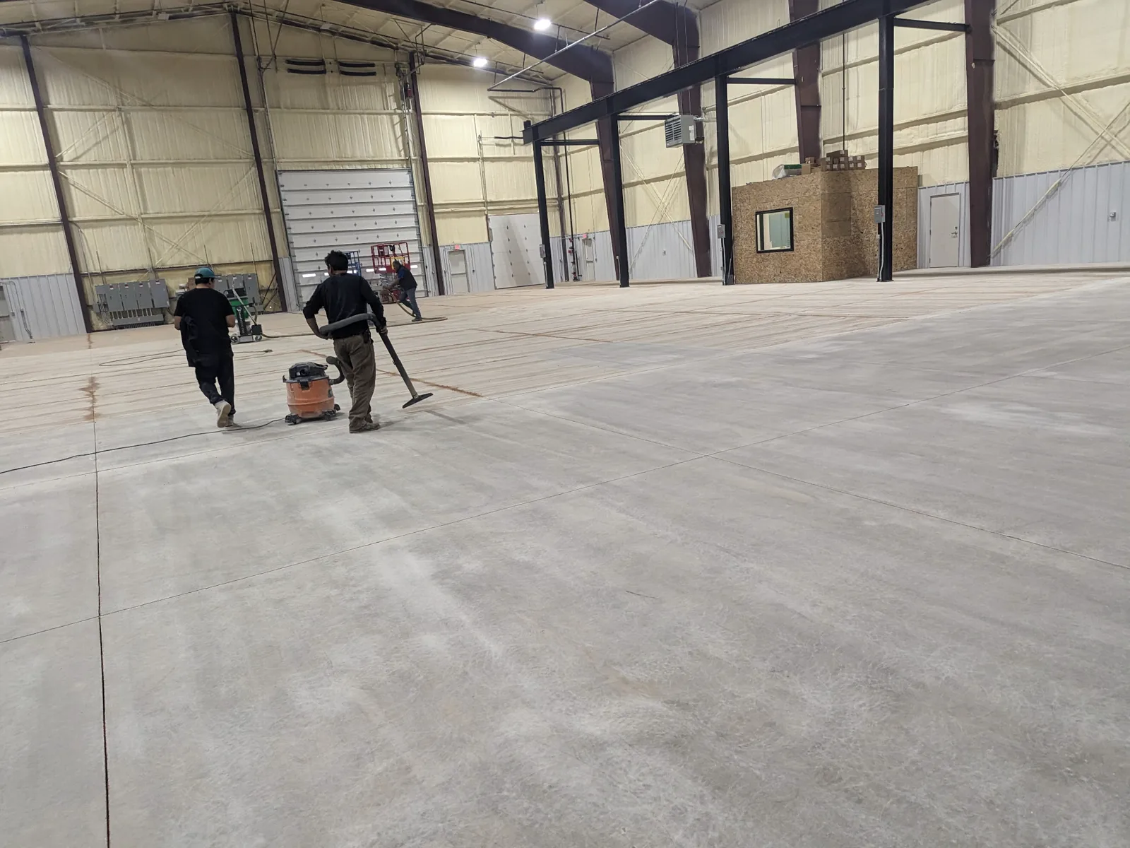 Two workers seen from behind vacuuming a large bare concrete slab inside a steel-framed building, with a shop vacuum and an extraction hose on the floor.
