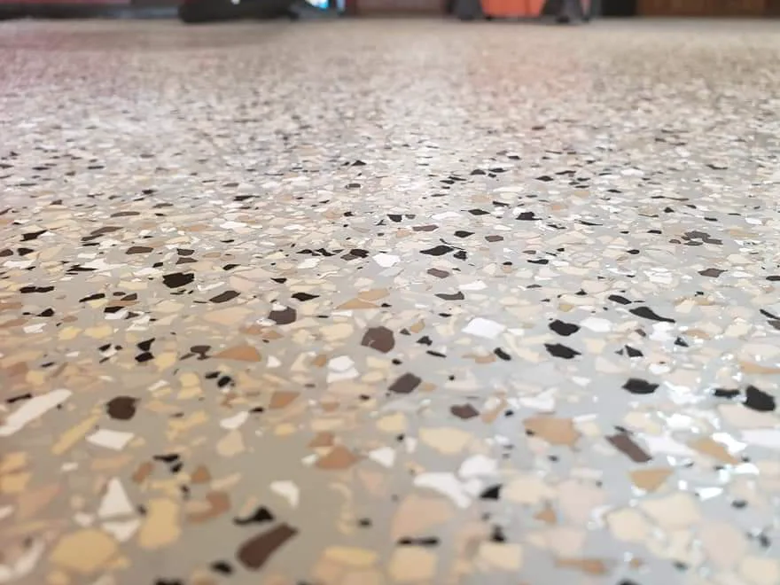 Close view of a floor surface showing individual cream, tan, brown and black decorative chips sealed beneath a glossy clear finish.