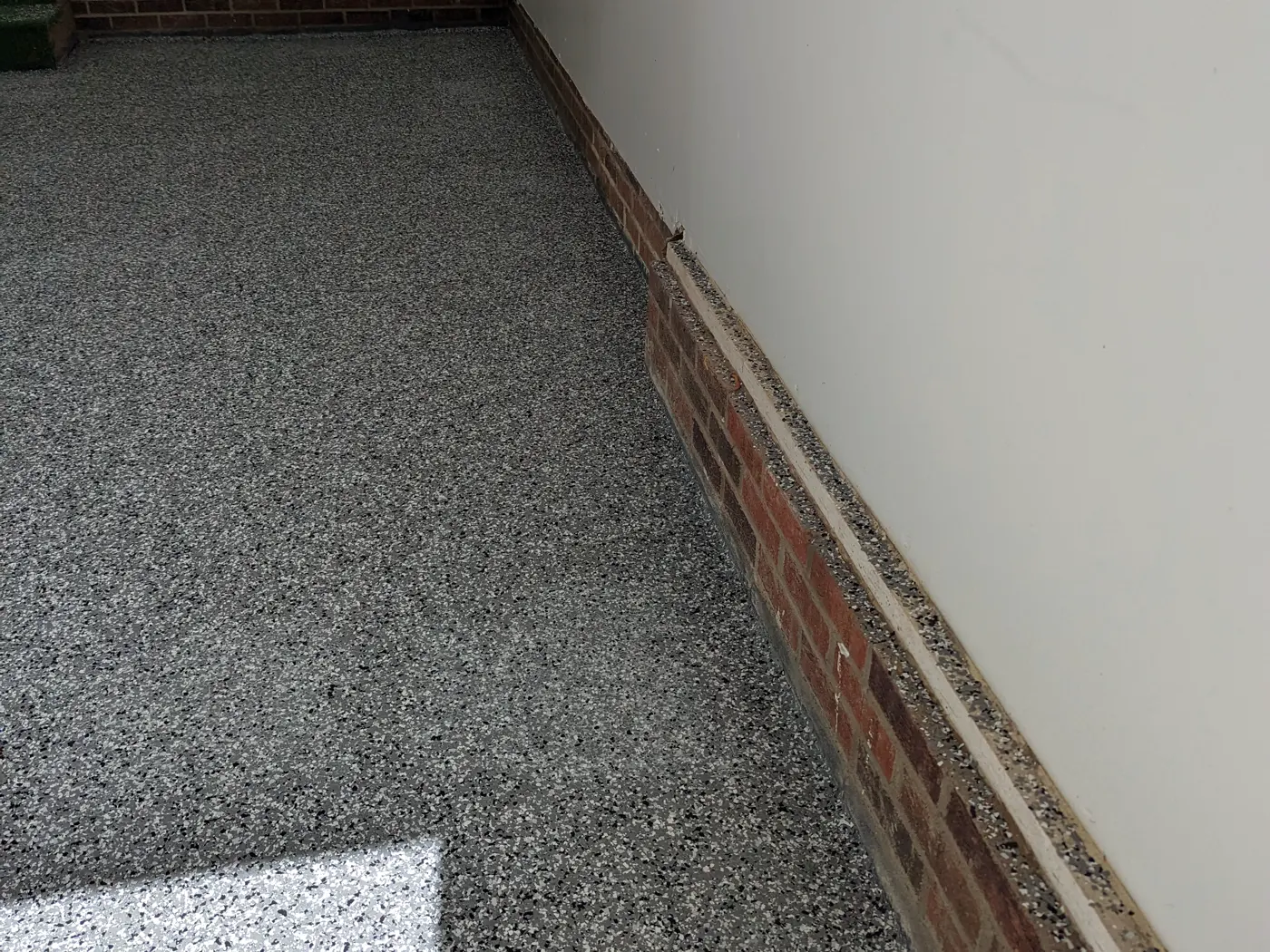 A grey decorative flake garage floor meeting a red brick stem wall, with the coating carried up over the top of the brick ledge and the painted wall above it left uncoated.