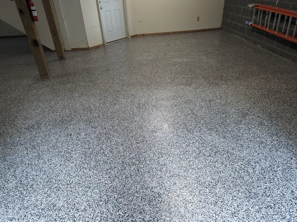 A finished garage floor in a warm grey and tan decorative flake blend, running past two timber posts towards a white door, with a dark block wall to the right.