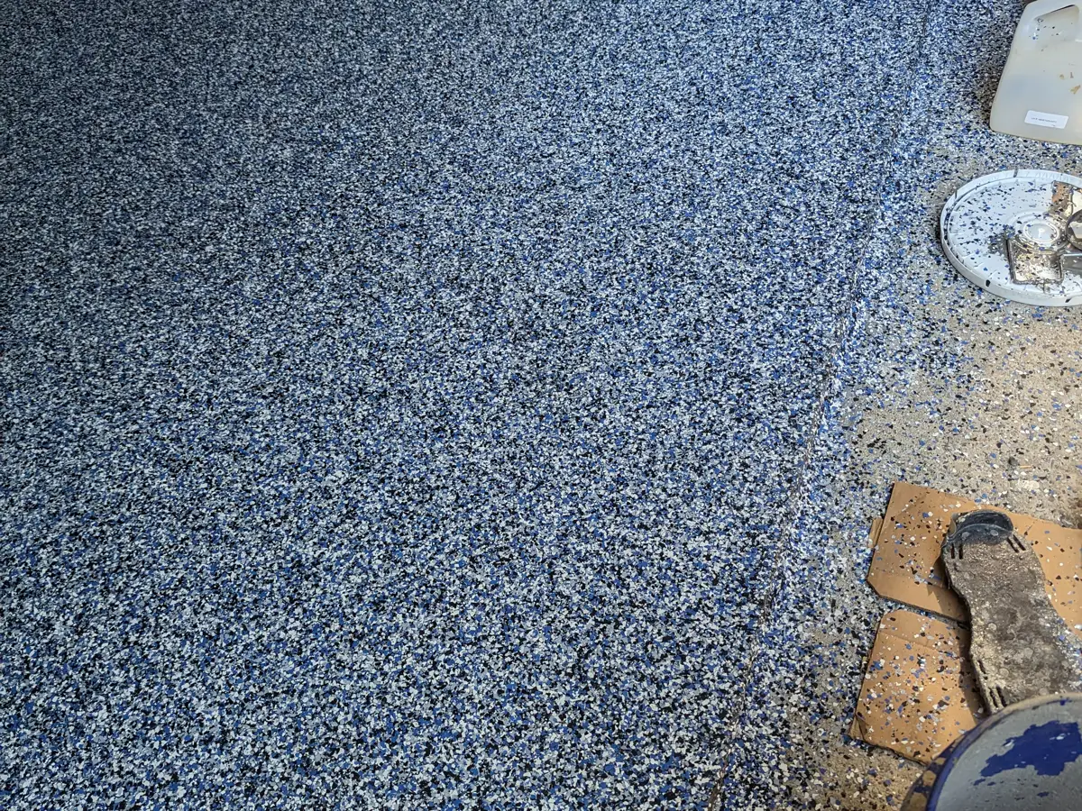A dense blue, white and black decorative broadcast covering most of a garage slab, with loose media scattered across the bare concrete still showing along the right-hand edge.