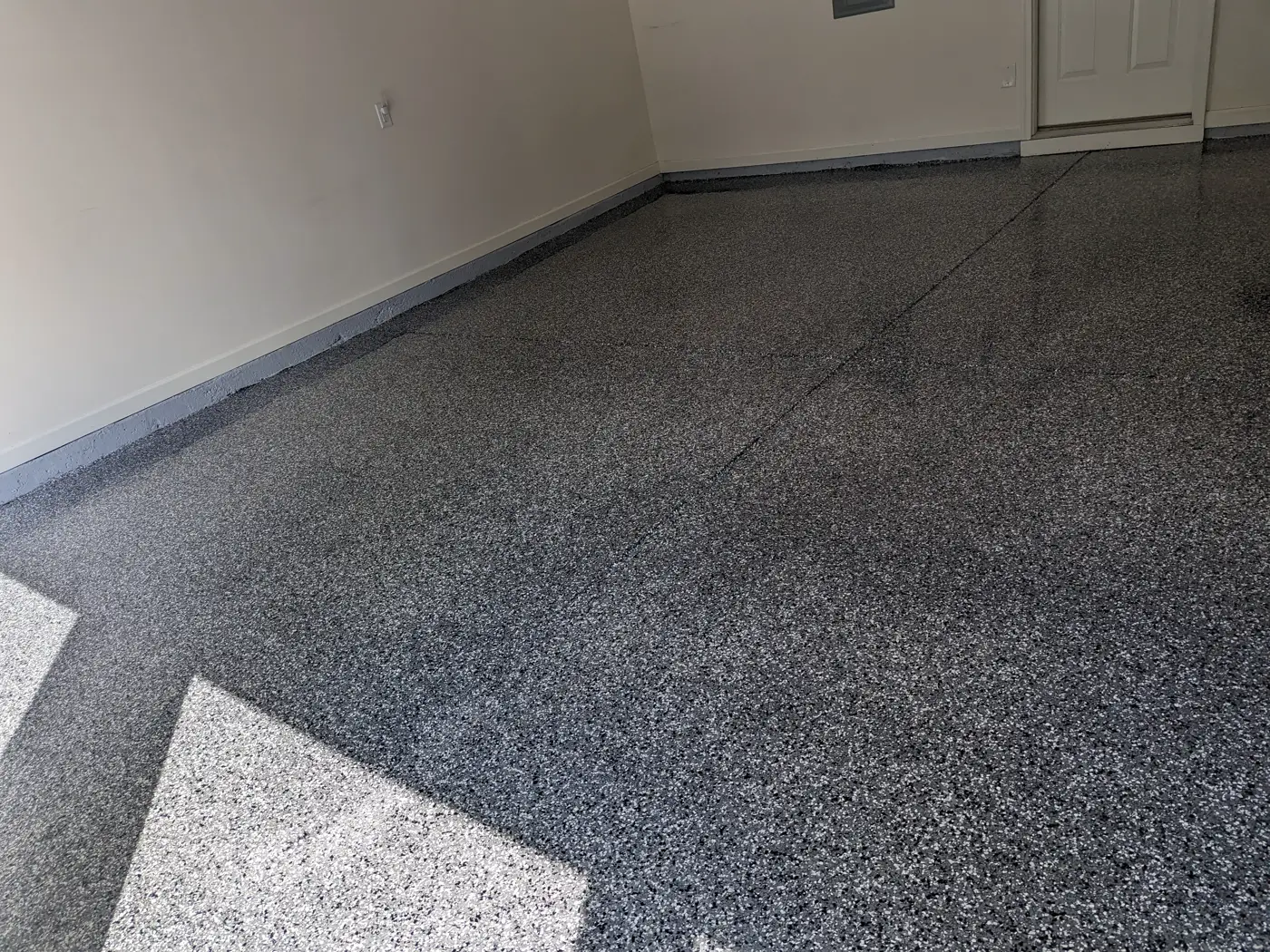A finished garage floor covered in dense grey, black and white decorative flake, running to a painted wall base and a white door, with the slab joints still visible through the surface.