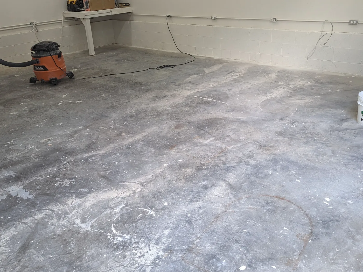 A bare garage slab before work starts, marked with paint spatter, dark staining and a rust ring, with a shop vacuum standing against the painted block wall.