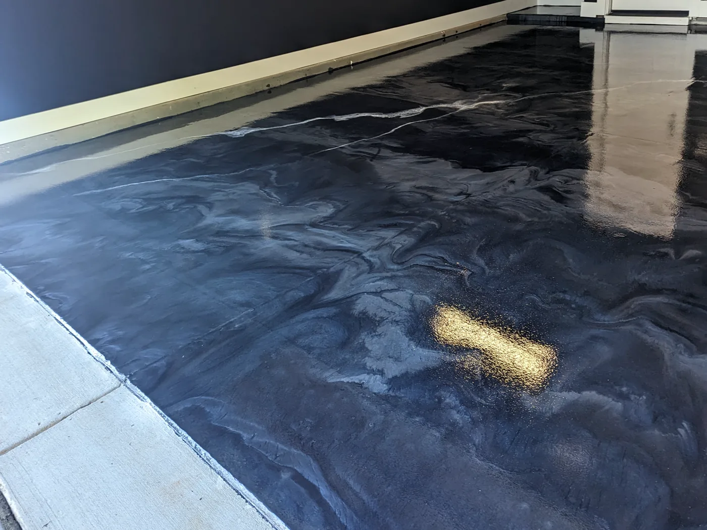 A dark blue-grey metallic floor in a garage, marbled with pale swirls and reflecting a doorway, meeting a strip of bare concrete along the garage door line.