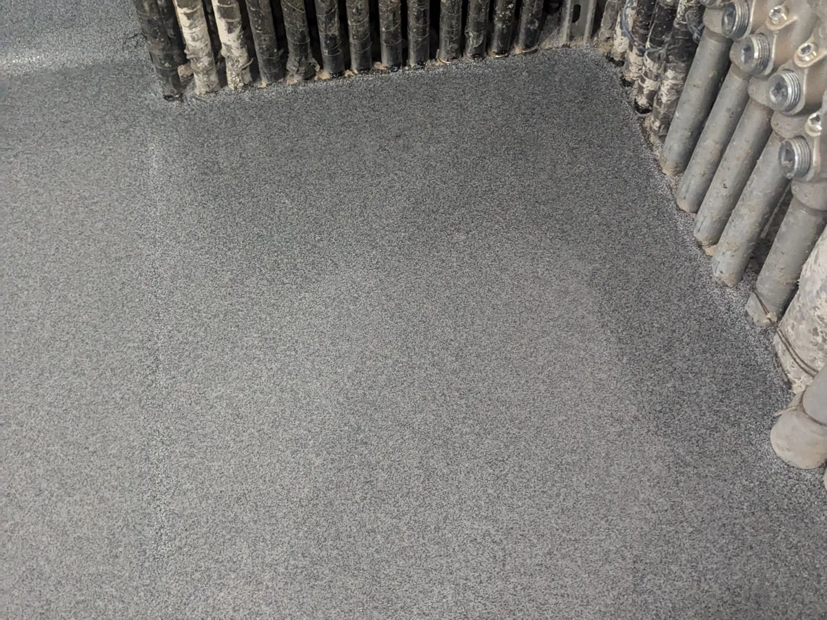 A pale grey textured floor coating worked closely around a dense bundle of conduits and pipes rising out of the slab, with the coating turning up the wall behind as a coved base.