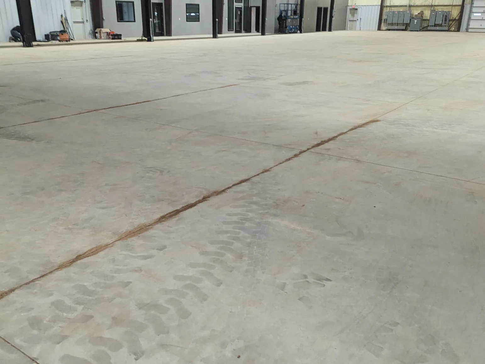 A large bare concrete slab inside a steel-framed industrial building, marked with equipment tire tracks and crossed by saw-cut joints, with electrical switchgear along the far wall.