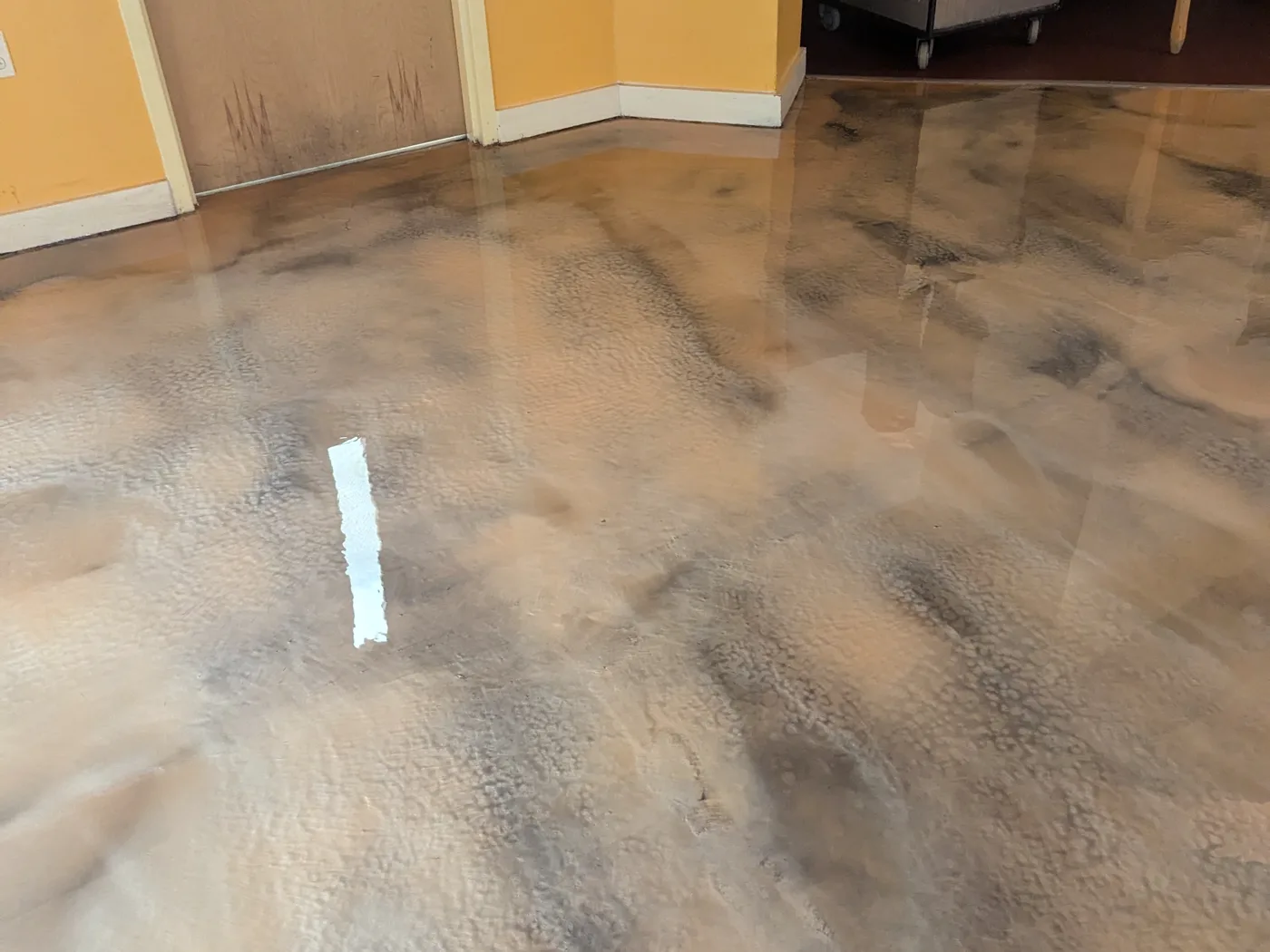 A copper and grey metallic floor in a working back-of-house area, running to a scuffed door and painted wall with a trolley parked at the edge of the frame.