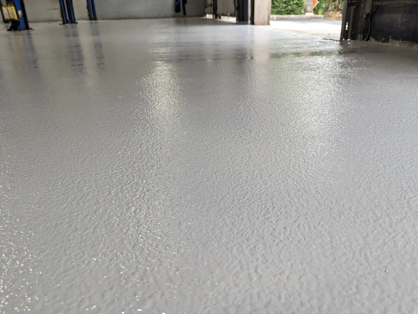 A low view along a pale grey coated floor in a vehicle service bay, the surface showing a fine even texture, with vehicle lifts to the left and an open bay door letting daylight in at the far end.