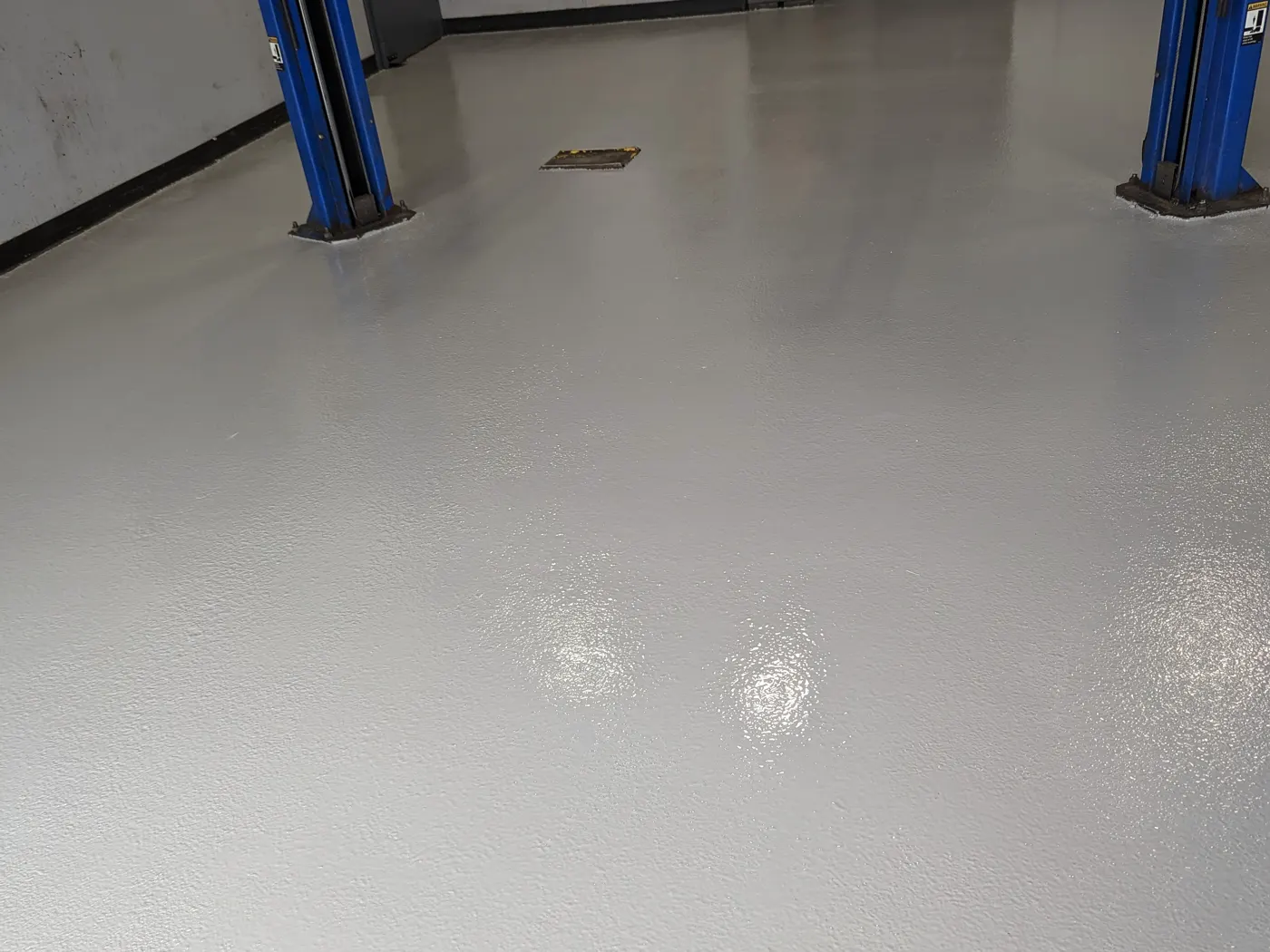 A freshly coated pale grey floor running between two blue vehicle lifts in a service bay, with a floor grate set into the surface and lights reflecting off the wet finish.